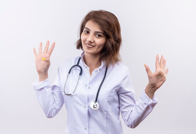 Young woman doctor in white coat with stethoscope raising hands in surrender smiling standing over white wall