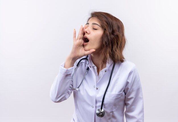 Young woman doctor in white coat with stethoscope shouting with hand near mouth standing over white wall