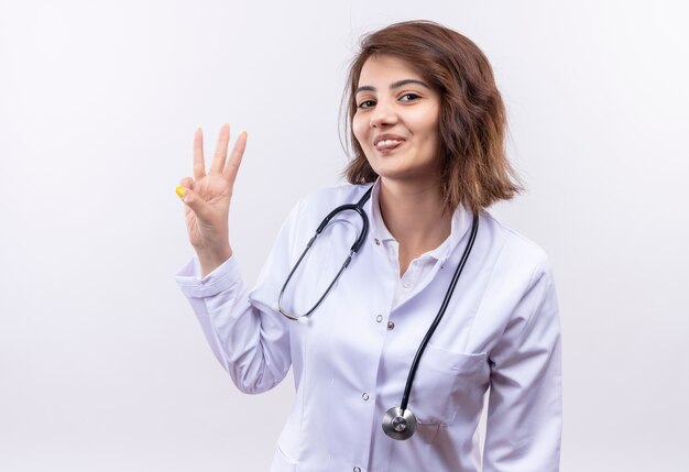 Young woman doctor in white coat with stethoscope showing and pointing up with fingers number three smiling standing over white wall