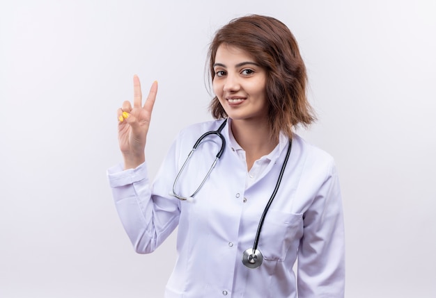 Young woman doctor in white coat with stethoscope showing and pointing up with fingers number two smiling standing over white wall