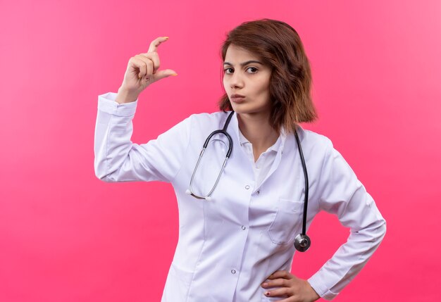 Young woman doctor in white coat with stethoscope showing small size sign with fingers, measure symbol standing over pink wall