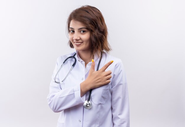 Молодая женщина-врач в белом халате со стетоскопом, весело улыбаясь, указывая указательным пальцем в сторону, стоящую над белой стеной