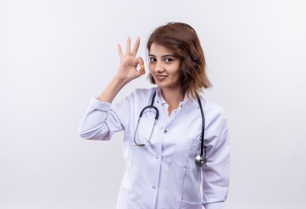 Young woman doctor in white coat with stethoscope smiling confident doing ok sign standing over white wall