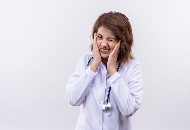 Young woman doctor in white coat with stethoscope touching face with hands with annoyed expression standing over white wall