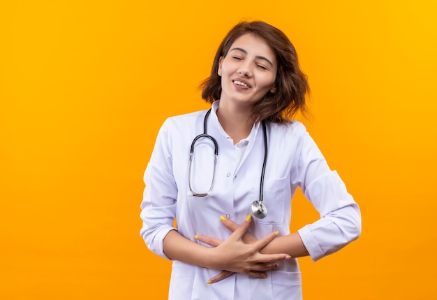 Young woman doctor in white coat with stethoscope with closed eyes touching her belly smiling standing over orange wall