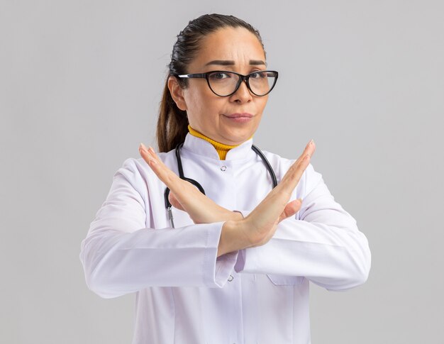 Молодая женщина-врач в белом медицинском халате в очках со стетоскопом на шее с серьезным лицом делает стоп-жест, скрещивая руки