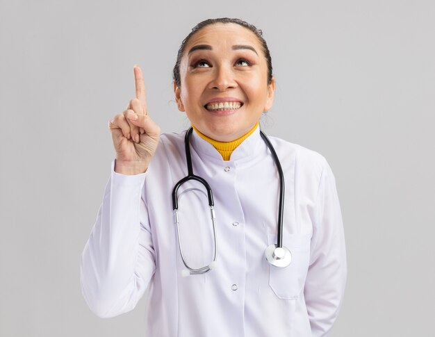Молодая женщина-врач в белом медицинском халате со стетоскопом на шее счастлива и радостно смотрит вверх, указывая указательным пальцем на что-то, стоящее над белой стеной