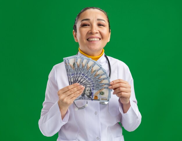 Young woman doctor in white medical coat with stethoscope around neck holding cash  with happy face standing over green wall