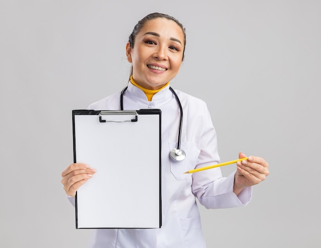 Молодая женщина-врач в белом медицинском халате со стетоскопом на шее держит буфер обмена с пустыми страницами и улыбается карандашом, прося подпись, стоя над белой стеной