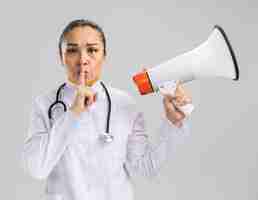 Free photo young woman doctor in white medical coat with stethoscope around neck holding megaphone  making silence gesture with finger on lips standing over white wall