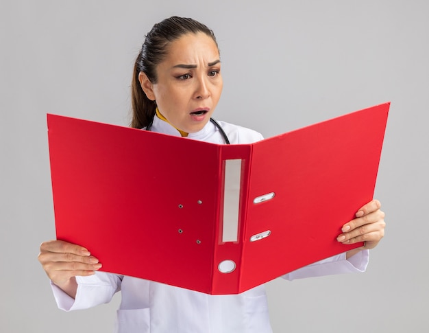 Молодая женщина-врач в белом медицинском халате со стетоскопом на шее держит красную папку, глядя на нее с недовольным лицом