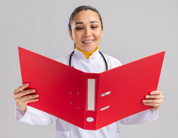 Young woman doctor in white medical coat with stethoscope around neck holding red folder looking at it with smile on face
