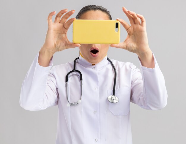 Young woman doctor in white medical coat with stethoscope around neck holding smartphone in front of her face being surprised