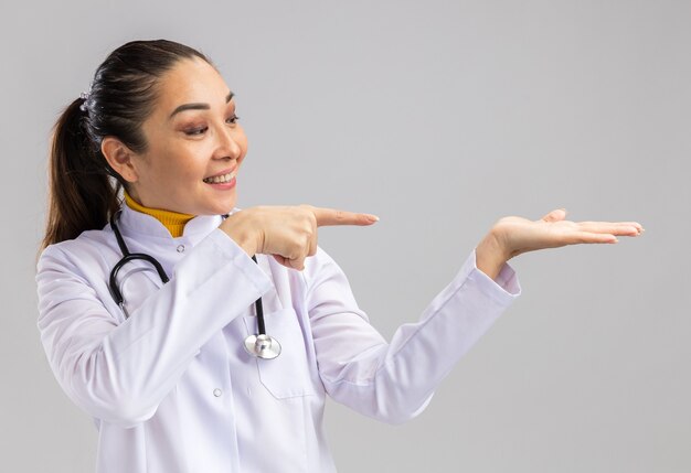 Young woman doctor in white medical coat with stethoscope around neck presenting with arm copy space pointing with index finger to the side standing over white wall