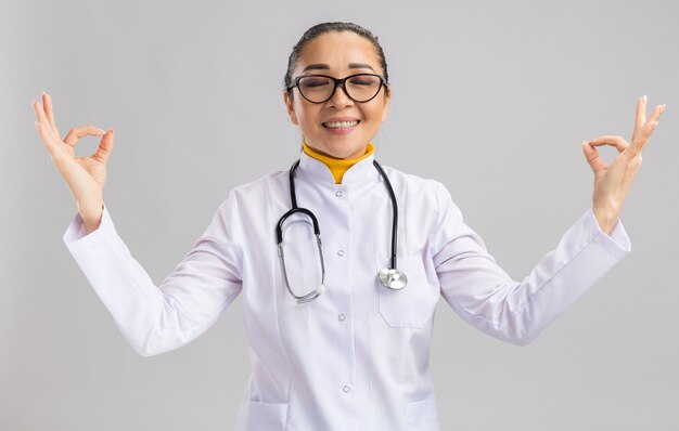 白い壁の上に立つ指で瞑想ジェスチャーを作るリラックスした首の周りに聴診器を持つ白い医療コートを着た若い女性医師