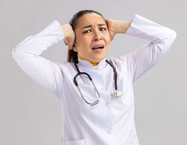 Young woman doctor in white medical coat with stethoscope around neck   with annoyed expression touching her head with hands