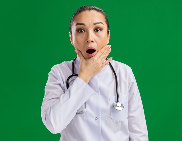 Young woman doctor in white medicine coat with stethoscope around neck  amazed and surprised standing over green wall