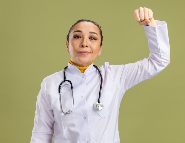 Medico della giovane donna in camice bianco della medicina con lo stetoscopio intorno al pugno serrato del collo felice e sicuro che sta sopra la parete verde