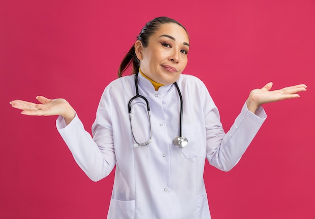 Medico della giovane donna in camice bianco della medicina con lo stetoscopio intorno al collo confuso che scrolla le spalle le spalle senza risposta in piedi sul muro rosa pink