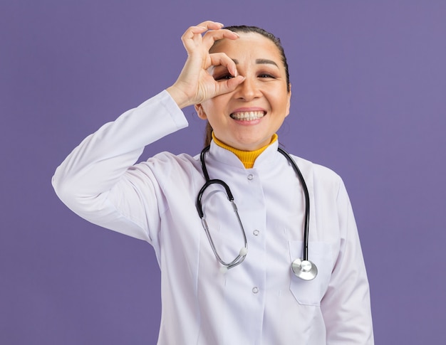 白い薬のコートを着た若い女性医師が、首に聴診器を付けて、紫色の壁の上に立って笑顔でこの看板を見て、okのサイン