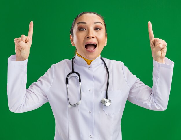목 주위에 청진기와 흰색 의학 코트에 젊은 여자 의사 녹색 벽 위에 서있는 새로운 아이디어를 갖는 행복하고 놀란 보여주는 인덱스 figners