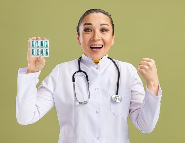 白い薬のコートを着た若い女性医師が、緑の壁の上に立って、幸せで興奮して拳を握りしめ、丸薬で水疱を首の周りに保持している