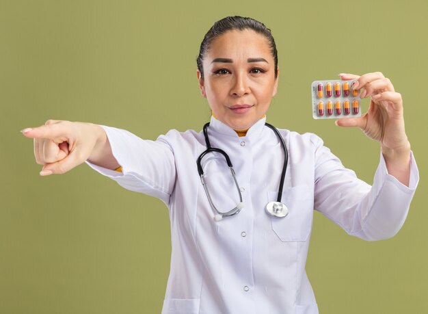 Medico della giovane donna in camice bianco della medicina con lo stetoscopio intorno al collo che tiene la bolla con le pillole che guarda l'obbiettivo con la faccia seria che indica con il dito indice a qualcosa sulla parete verde