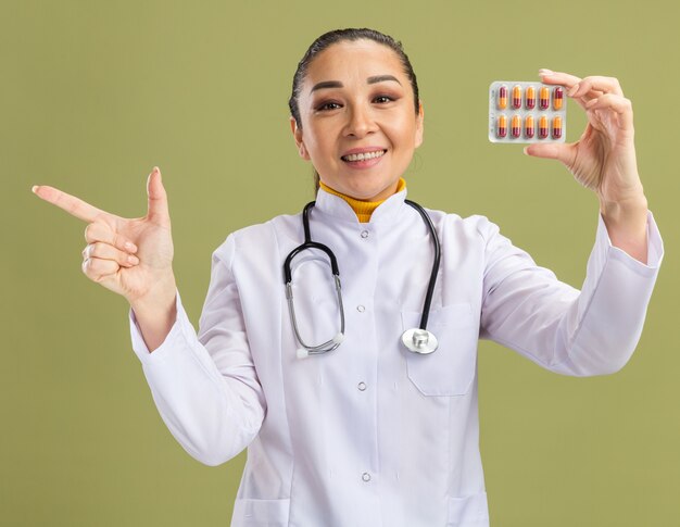 Foto gratuita medico della giovane donna in camice bianco della medicina con lo stetoscopio intorno al collo che tiene il blister con le pillole che guarda l'obbiettivo con il sorriso sul fronte che indica con il dito indice al lato sulla parete verde