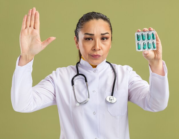 Молодая женщина-врач в белом халате со стетоскопом на шее держит блистер с таблетками со скептическим выражением лица, делая стоп-жест рукой, стоящей над зеленой стеной