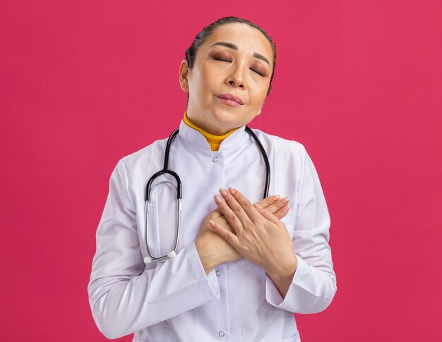 Young woman doctor in white medicine coat with stethoscope around neck  holding hands on chest feeling thankful standing over pink wall