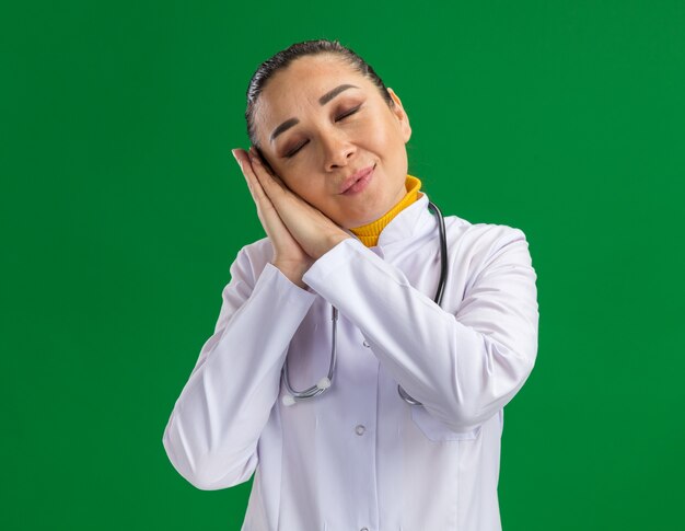 Young woman doctor in white medicine coat with stethoscope around neck holding palms together making sleep gesture leaning head on palms standing over green wall