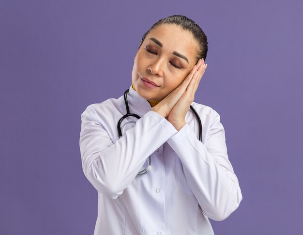 Young woman doctor in white medicine coat with stethoscope around neck holding palms together making sleeping gesture leaanung head on palms standing over purple wall