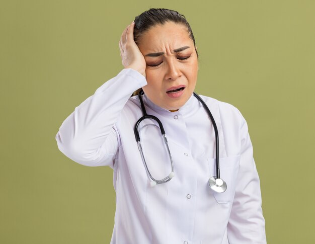 Young woman doctor in white medicine coat with stethoscope around neck looking unwell touching her head suffering from headache standing over green wall
