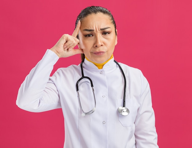 Young woman doctor in white medicine coat with stethoscope around neck pointing with index finger at her temple  with skeptic expression standing over pink wall