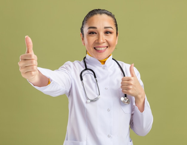 Молодая женщина-врач в белом халате со стетоскопом на шее, уверенно улыбаясь, показывает палец вверх, стоя над зеленой стеной