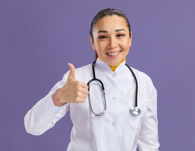 Молодая женщина-врач в белом халате со стетоскопом на шее, уверенно улыбаясь, показывает палец вверх, стоя над фиолетовой стеной