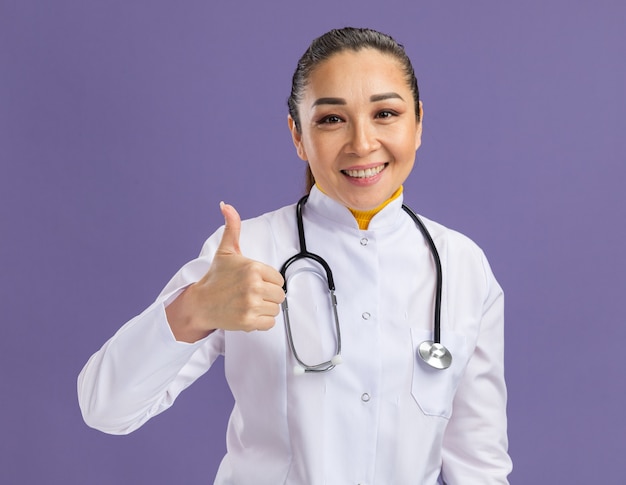 Young woman doctor in white medicine coat with stethoscope around neck  smiling confident showing thumbs up standing over purple wall