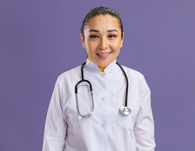 Молодая женщина-врач в белом халате со стетоскопом на шее, уверенно улыбаясь, стоя над фиолетовой стеной