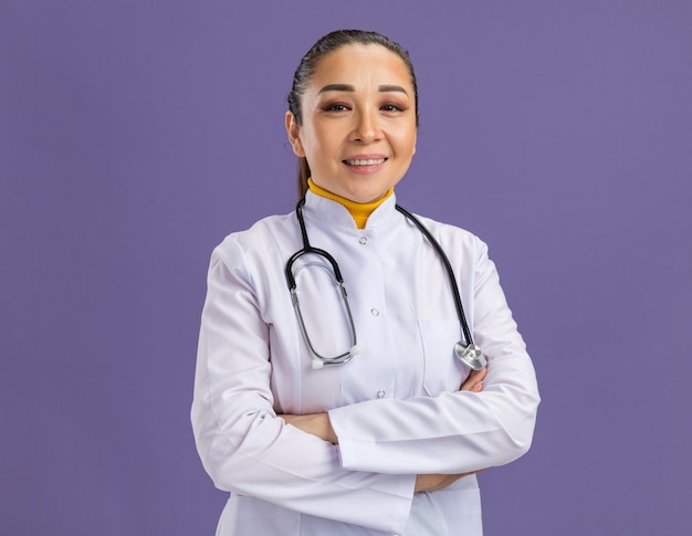 Young woman doctor in white medicine coat with stethoscope around neck  smiling confident with arms crossed standing over purple wall