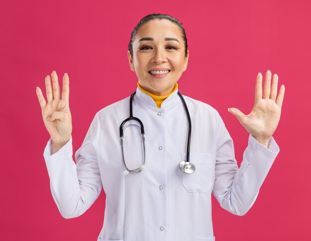 Young woman doctor in white medicine coat with stethoscope around neck  smiling showing number nine with fingers standing over pink wall