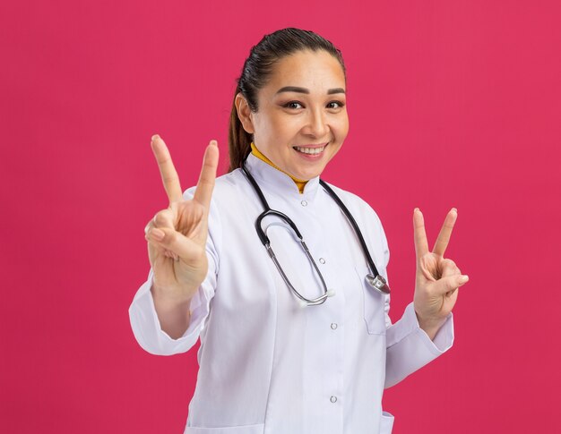 Medico della giovane donna in camice bianco della medicina con lo stetoscopio intorno al collo che sorride mostrando il v-sign che sta sopra la parete rosa