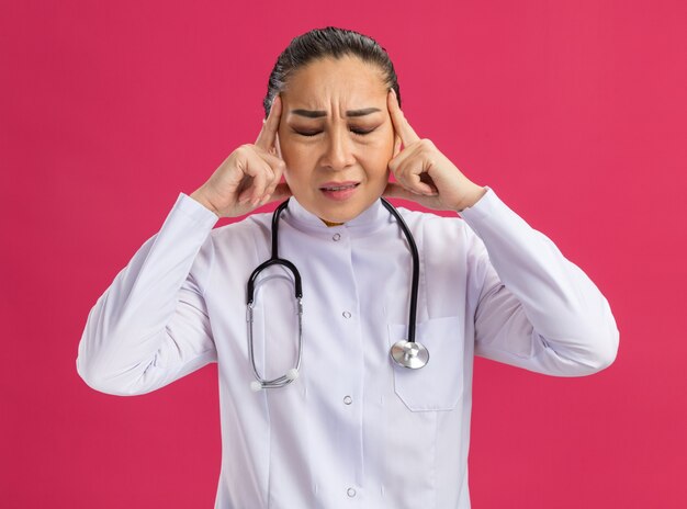 Young woman doctor in white medicine coat with stethoscope around neck touching her temples with annoyed expression suffering from headache standing over pink wall