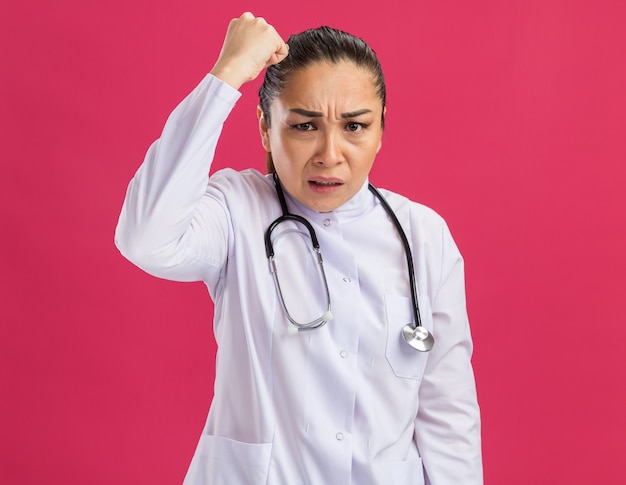 Free photo young woman doctor in white medicine coat with stethoscope around neck  with angry face showing fist standing over pink wall