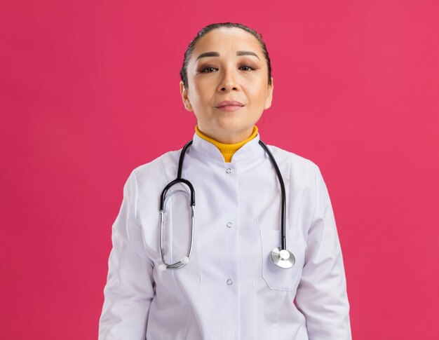 ピンクの壁の上に立つ自信のある表情で聴診器を首にかけた白い薬のコートを着た若い女性医師
