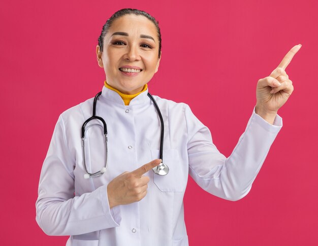 분홍색 벽 위에 서있는 측면에 검지 손가락으로 가리키는 얼굴에 미소로 목 주위에 청진기와 흰색 의학 코트에 젊은 여자 의사