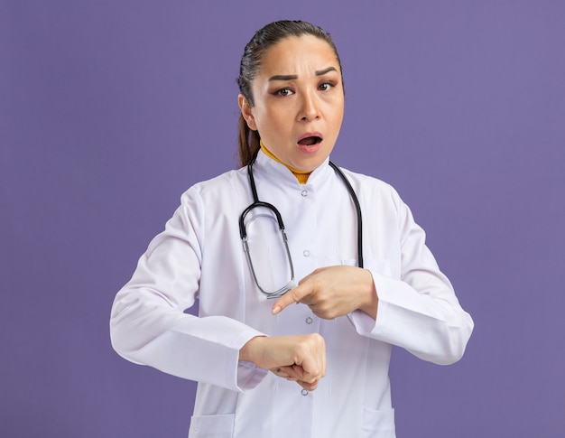 Medico della giovane donna in camice bianco della medicina con lo stetoscopio intorno al collo con l'espressione preoccupata che indica il suo braccio che ricorda il tempo che sta sopra la parete viola