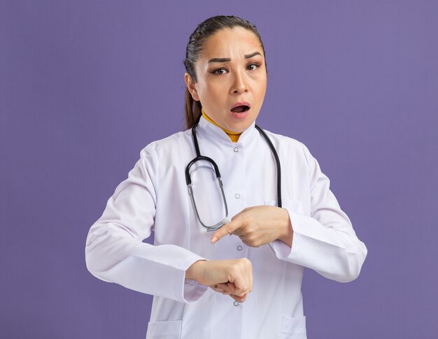 Young woman doctor in white medicine coat with stethoscope around neck  with worried expression pointing at her arm reminding about time standing over purple wall