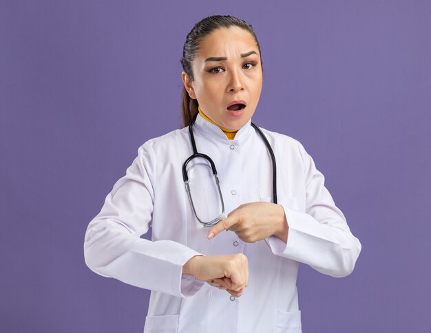 白い薬のコートを着た若い女性医師で、聴診器を首にかけ、心配そうな表情で腕を指し、紫色の壁の上に立つ時間を思い出させる