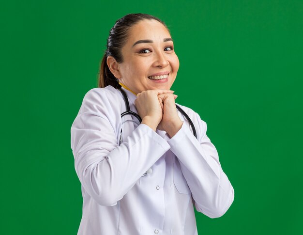 Young woman doctor     with shy smile on face waiting for surprise holding hands together