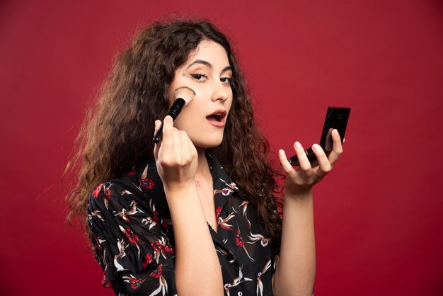 Young woman doing makeup on a red wall.